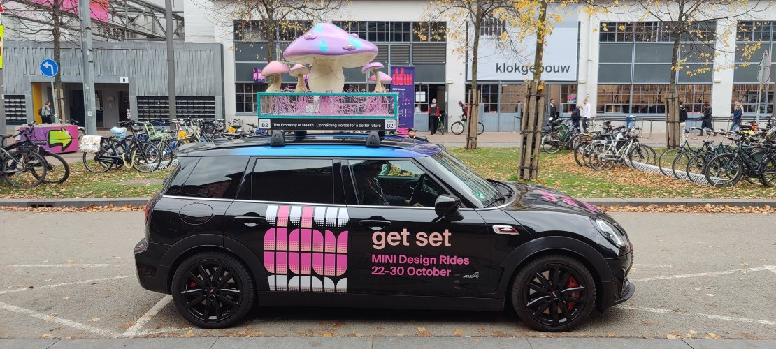 A mycelium-inspired installation featuring large purple mushrooms with turquoise details mounted on a transparent base filled with illuminated strands, displayed on the roof of a Mini Ride car during Dutch Design Week 2022.